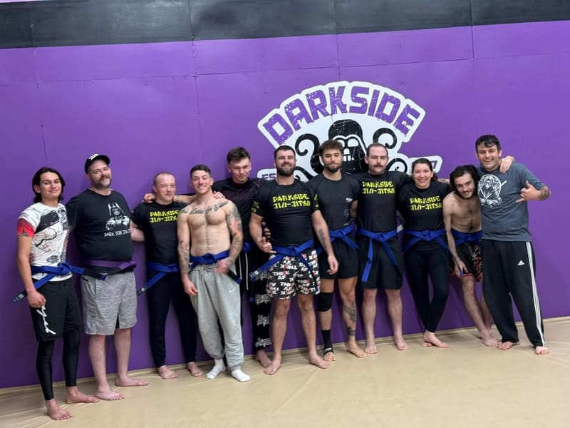 A group of Dark Side students with brand-new belts, lined up in front of the purple Darkside logo wall on promotion day